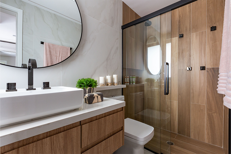 Projeto: Daniela Funari| No banheiro master, materiais naturais criam clima de spa. O espelho redondo iluminado se destaca, e o porcelanato marmorizado off white nas paredes harmoniza com o amadeirado do box.