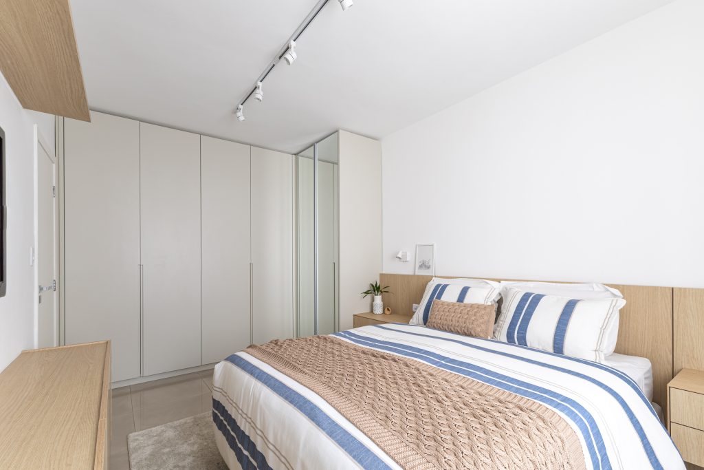 Projeto: Bima Arquitetura | No quarto de casal, marcenaria clara em L e cabeceira de madeira com iluminação embutida unem conforto e funcionalidade.