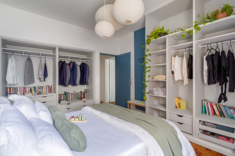 Projeto: Julia Otaga | No quarto do casal, o armário aberto em marcenaria sob medida organiza roupas, livros e objetos pessoais de forma prática e acolhedora. A pintura azul destaca portas e paredes, enquanto o uso de plantas traz frescor e leveza ao ambiente.