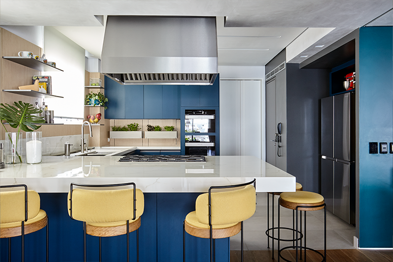 Projeto: Juliana Fabrizzi | A cozinha integrada combina armários azul-marinho, bancada em mármore e detalhes em madeira, criando um espaço moderno e acolhedor.