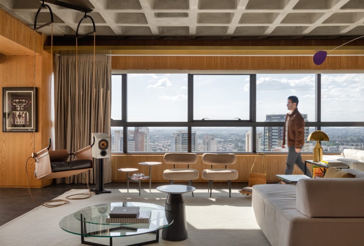 Projeto: Rodolfo Fontana | A área social do Apartamento Mike combina a imponência da laje aparente com mobiliário contemporâneo e vistas panorâmicas da cidade, em um espaço que equilibra linguagem industrial e conforto