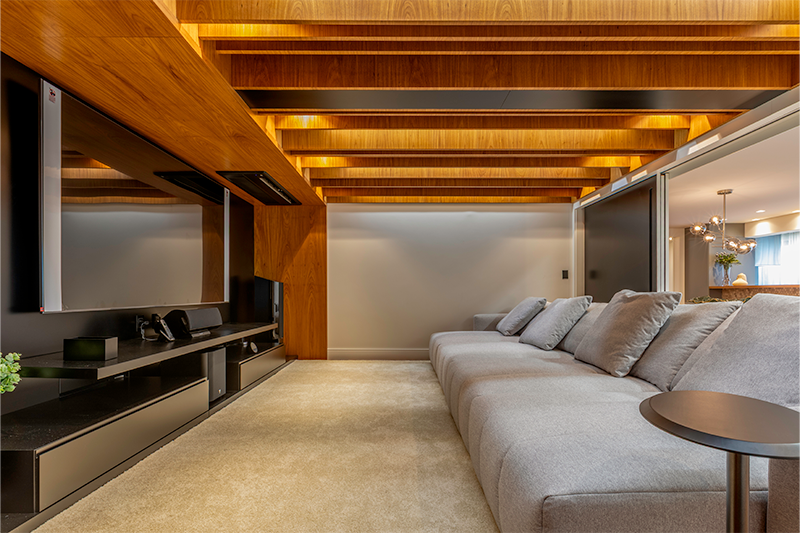 O home theater ganhou um teto com vigas de madeira em alturas variadas, criado a partir da necessidade de integrar uma viga estrutural existente.