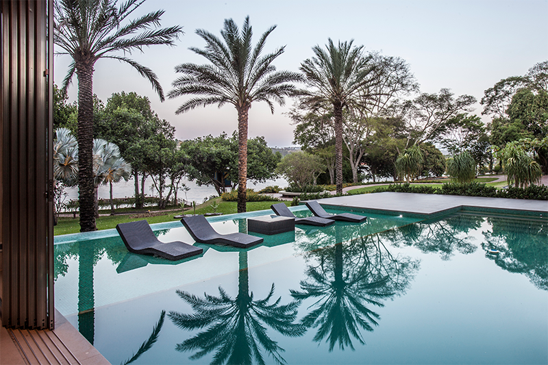 Projeto de paisagismo: Depieri Paisagismo|Na composição do paisagismo desta residência localizada em Brasília, os arredores da piscina revelam um amplo gramado verde e espécies como palmeiras, árvores, forrações e arbustos.