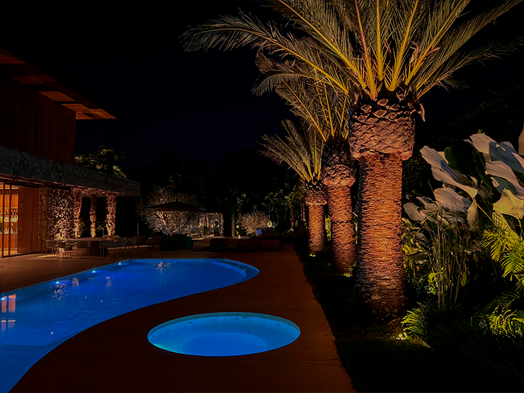 Projeto: MAAI Arquitetura |Mais um exemplo de jardim com vegetação tropical e palmeiras do tipo Canariense. A imagem noturna do projeto traz a visão da piscina iluminada, cenário ideal para festas e encontros especiais.