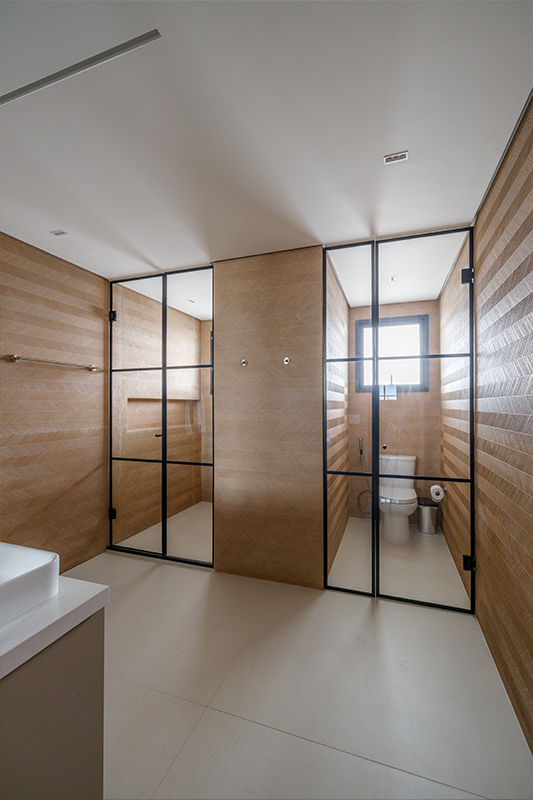 Projeto: Yannick Athia| Banheiro da suíte principal revestido do piso ao teto, com duas cubas, chuveiro e bacia sanitária em cabines separadas, garantindo conforto e funcionalidade.
