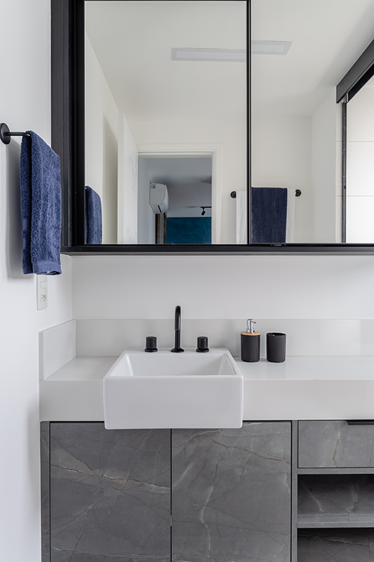 Projeto: Camila Palladino| No banheiro da suíte, a profissional utilizou tons neutros e uma marcenaria com aspecto de pedra cinza. Os metais pretos reforçam a estética urbana do projeto, enquanto o espelho amplo e o box em vidro minimalista garantem leveza e amplitude ao espaço.