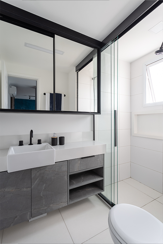 Projeto: Camila Palladino| No banheiro da suíte, a profissional utilizou tons neutros e uma marcenaria com aspecto de pedra cinza. Os metais pretos reforçam a estética urbana do projeto, enquanto o espelho amplo e o box em vidro minimalista garantem leveza e amplitude ao espaço.