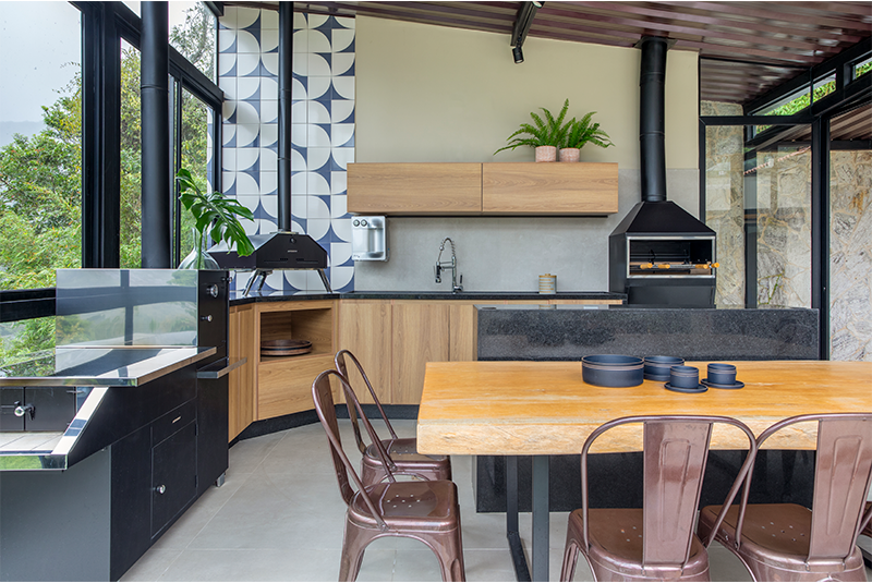 Projeto: Felipe Rohen Arquitetura | Fogão à lenha, forno de pizza e churrasqueira surgem em versões inox industriais, contrastando com marcenaria em freijó e mesa de tronco natural.